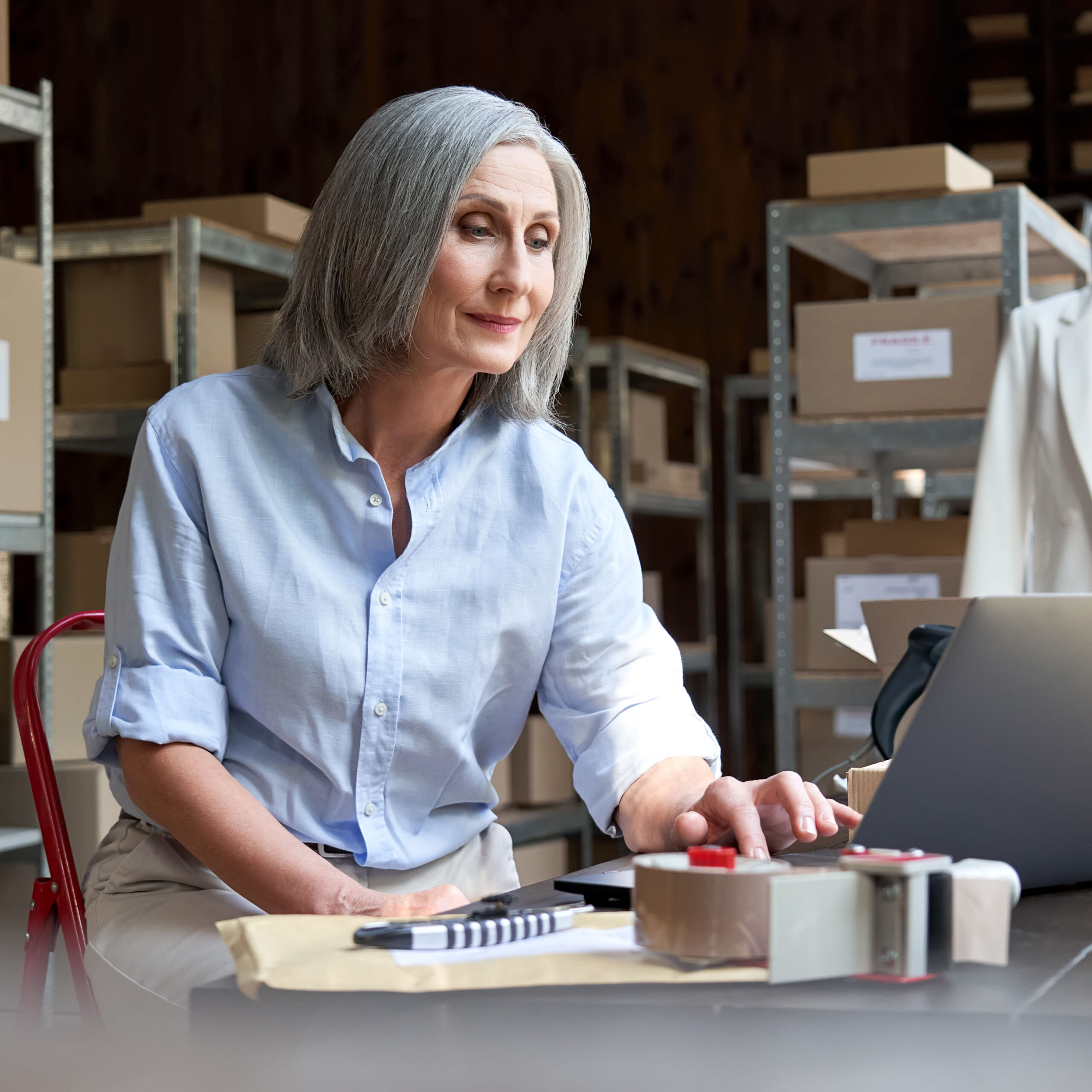 Mature female fashion seller using computer checking ecommerce clothing store orders. Older middle aged business woman entrepreneur working on laptop preparing online shipping delivery parcels boxes.
