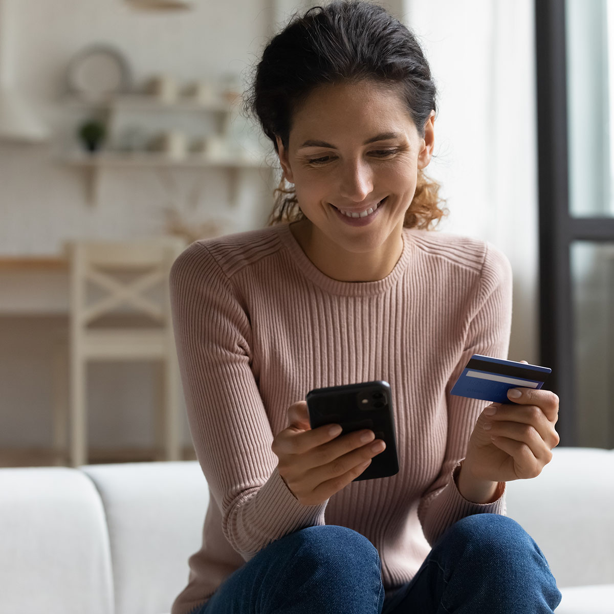 A slender woman with long curly hair in a ponytail, wearing a sweater and jeans, smiles at her phone while she holds a credit card in her other hand.