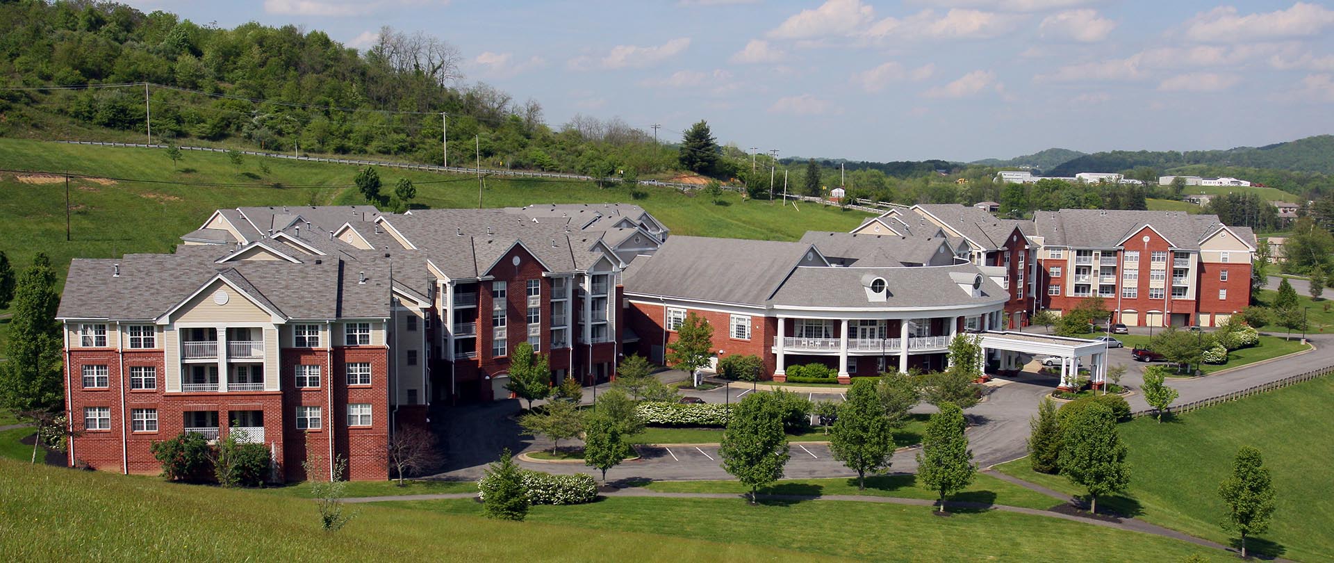 A wide angle photo of a red brock retirement community building in a hilly area surrounded by green landscape