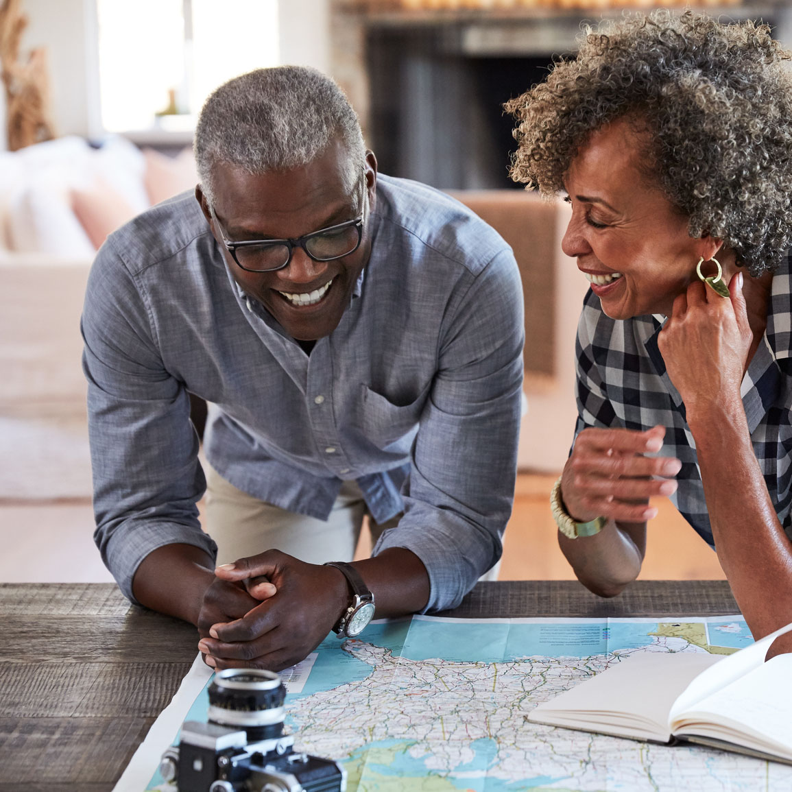 An older couple, a black man and a black woman, laugh with each other while leaning over a map on a table. It appears they are planning a trip.
