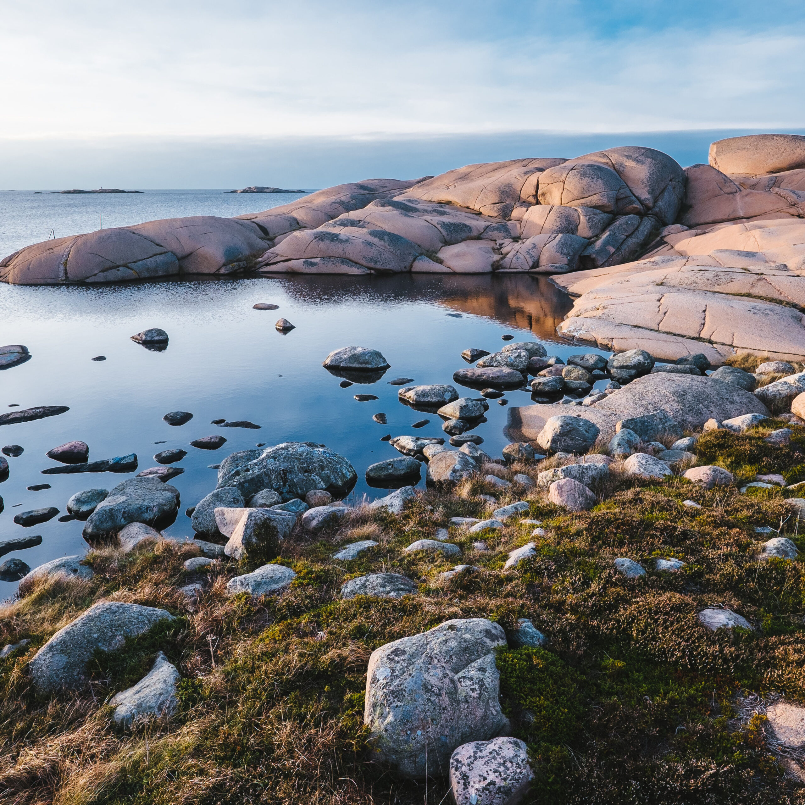 Photo of large boulders and smooth rocks at the edge of a body of water. The water is smooth and reflective and the grass on the edge of the short is green.