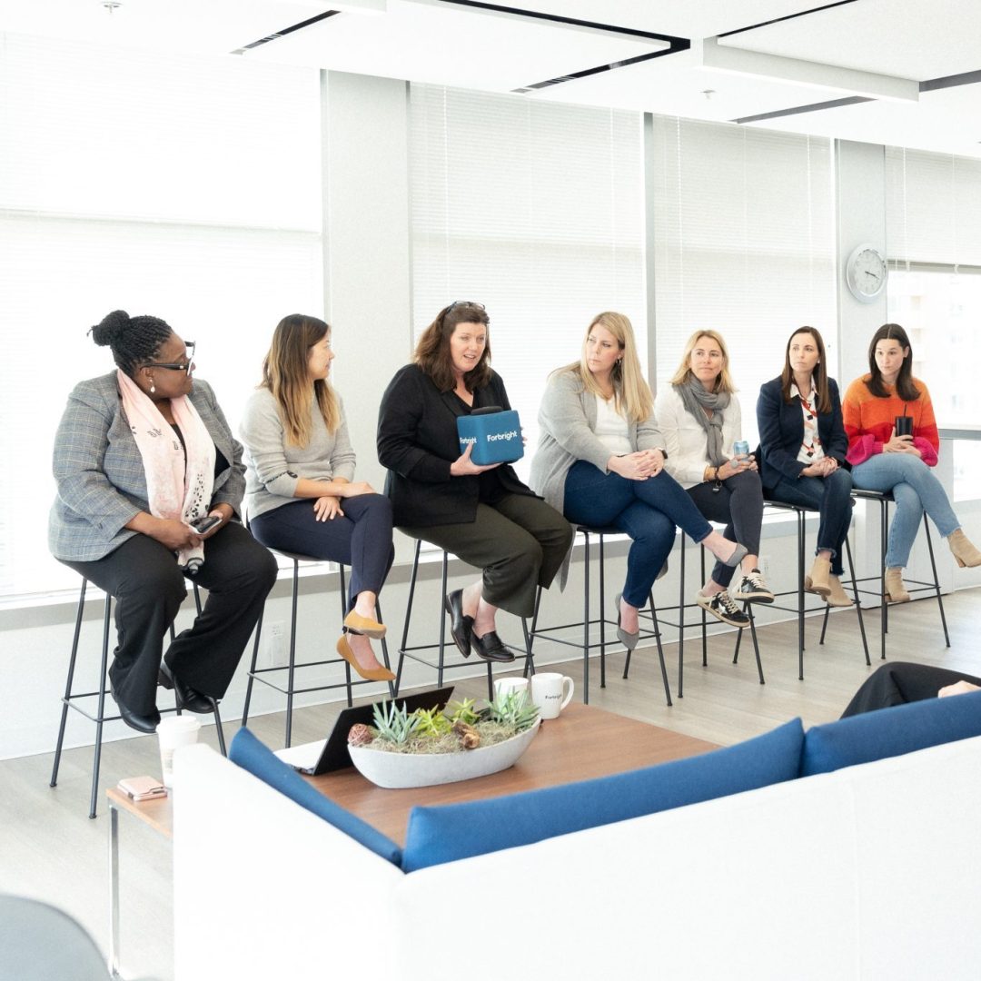 A panel of women sitting in chairs against a wall speaking during an event. The group is diverse in age and race.