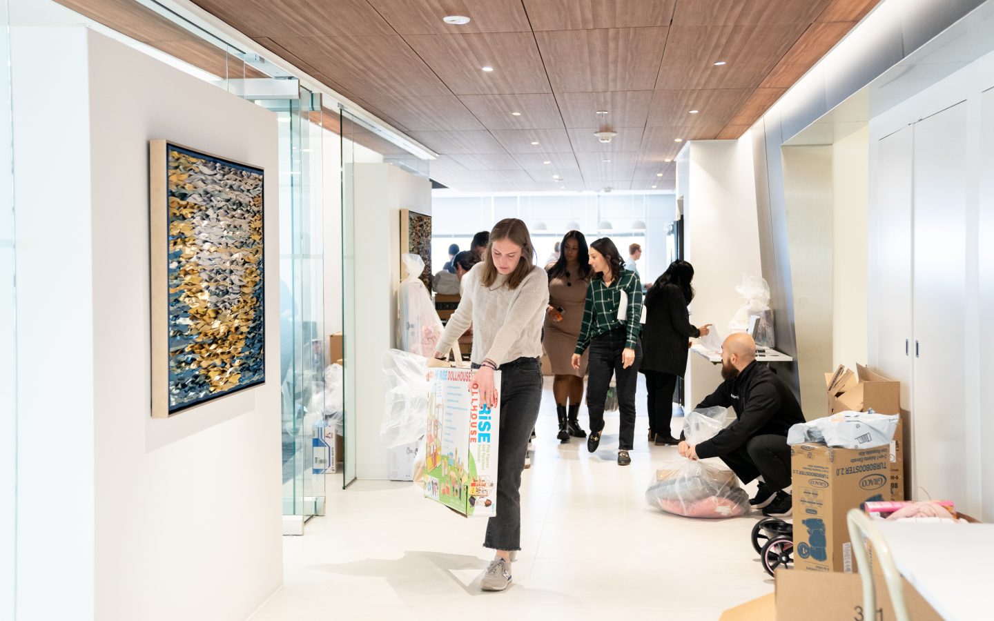 A woman walks towards the camera carrying a large dollhouse box while employees wrap and carry gifts around the Forbright Bank headquarters office for the Salvation Army Angel Tree program.