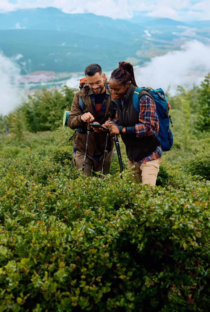 A Hispanic man and a black woman standing in shrubbery dressed in hiking gear. They are on a mountain, and behind them is a valley.
