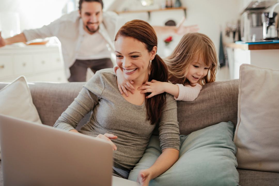 A white woman smiling as she studies her laptop on a sofa, while her playful partner and young daughter have fun in the background.