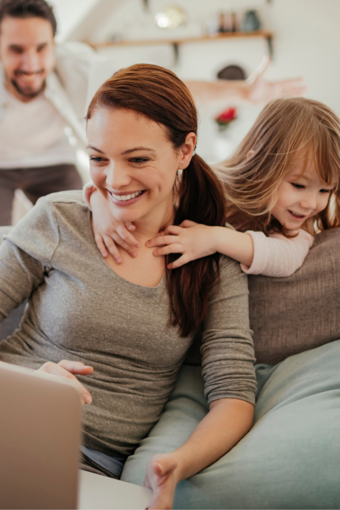 A white woman smiling as she studies her laptop on a sofa, while her playful partner and young daughter have fun in the background.