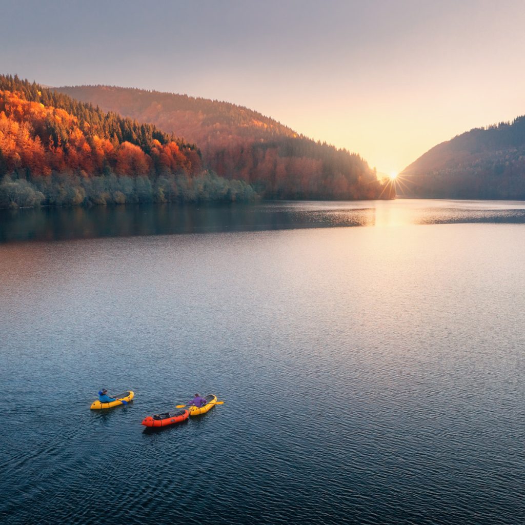 Aerial view of people on floating kayaks on blue lake with autumn mountains and sunset in distance.