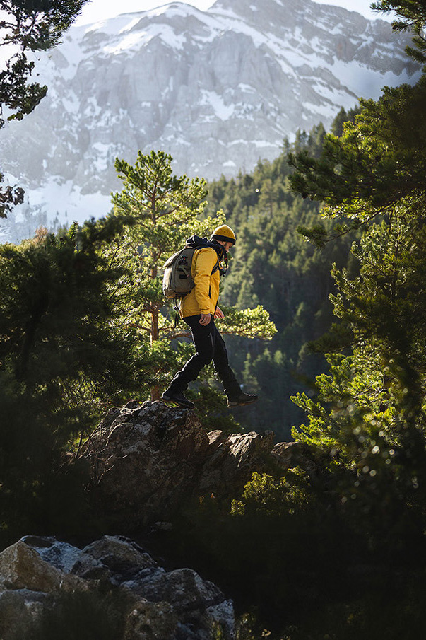 A man in hiking attire steps from one rock to the next as he explores a mountain. The sun is high, and behind him are lush trees.