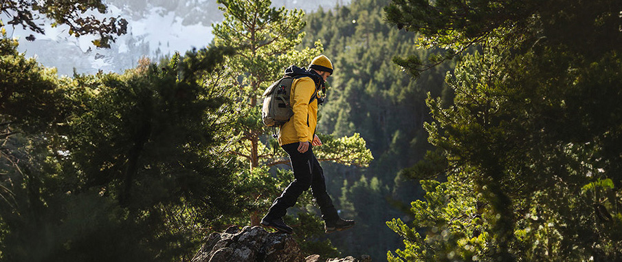 A man in hiking attire steps from one rock to the next as he explores a mountain. The sun is high, and behind him are lush trees.