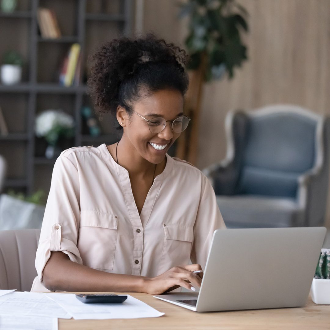 Young black woman smiling while working on laptop computer. Her hair is pulled back in a ponytail and she is wearing a pale pink shirt.