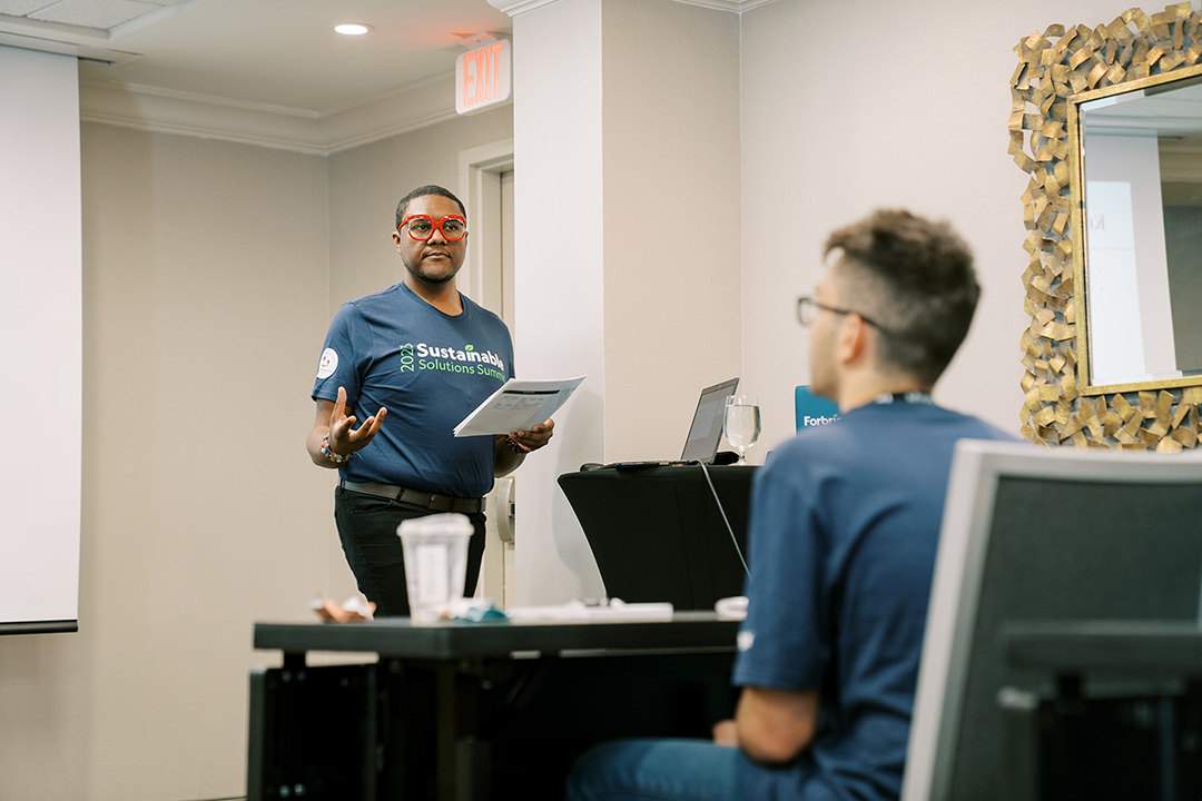 A Forbright employee wearing a Forbright T-shirt speaks to a group of students. He is a black man with short hair and is wearing red glasses.
