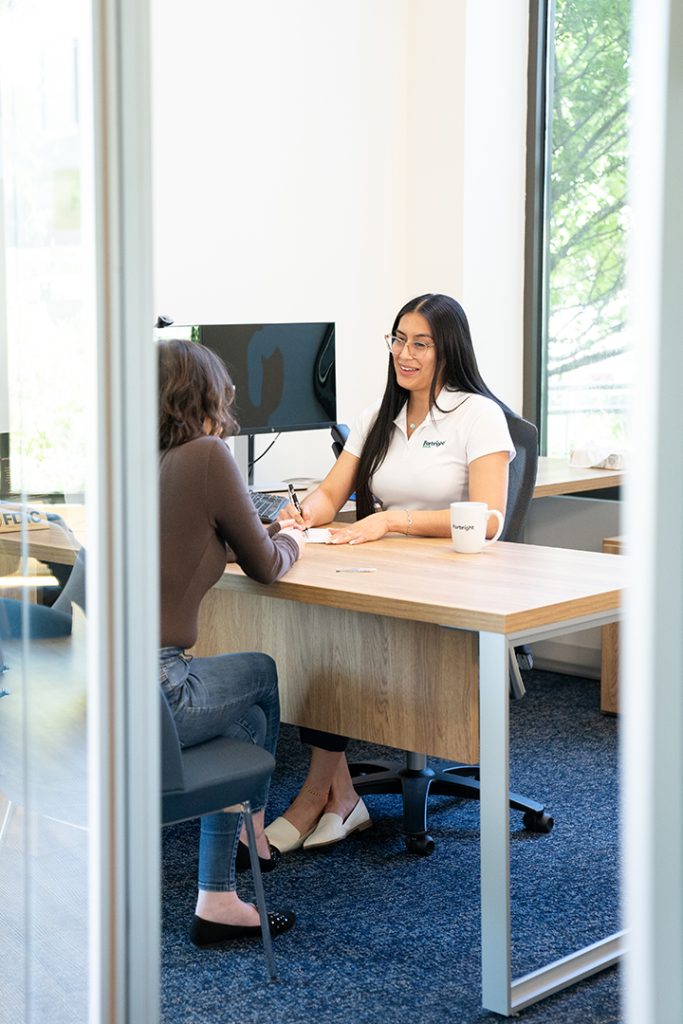 A female Forbright employee with long dark hair, wearing a white Forbright polo, assists a female customer while they sit at a desk in a Forbright Service Hub