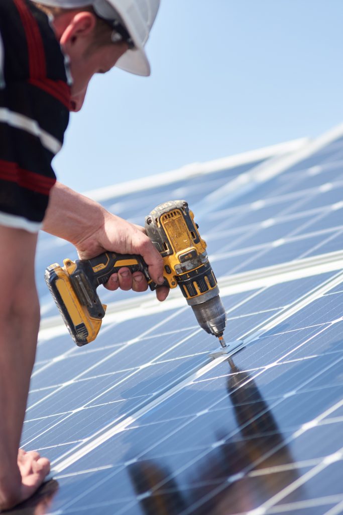 Male engineer in protective helmet installing solar photovoltaic panel system using drill.