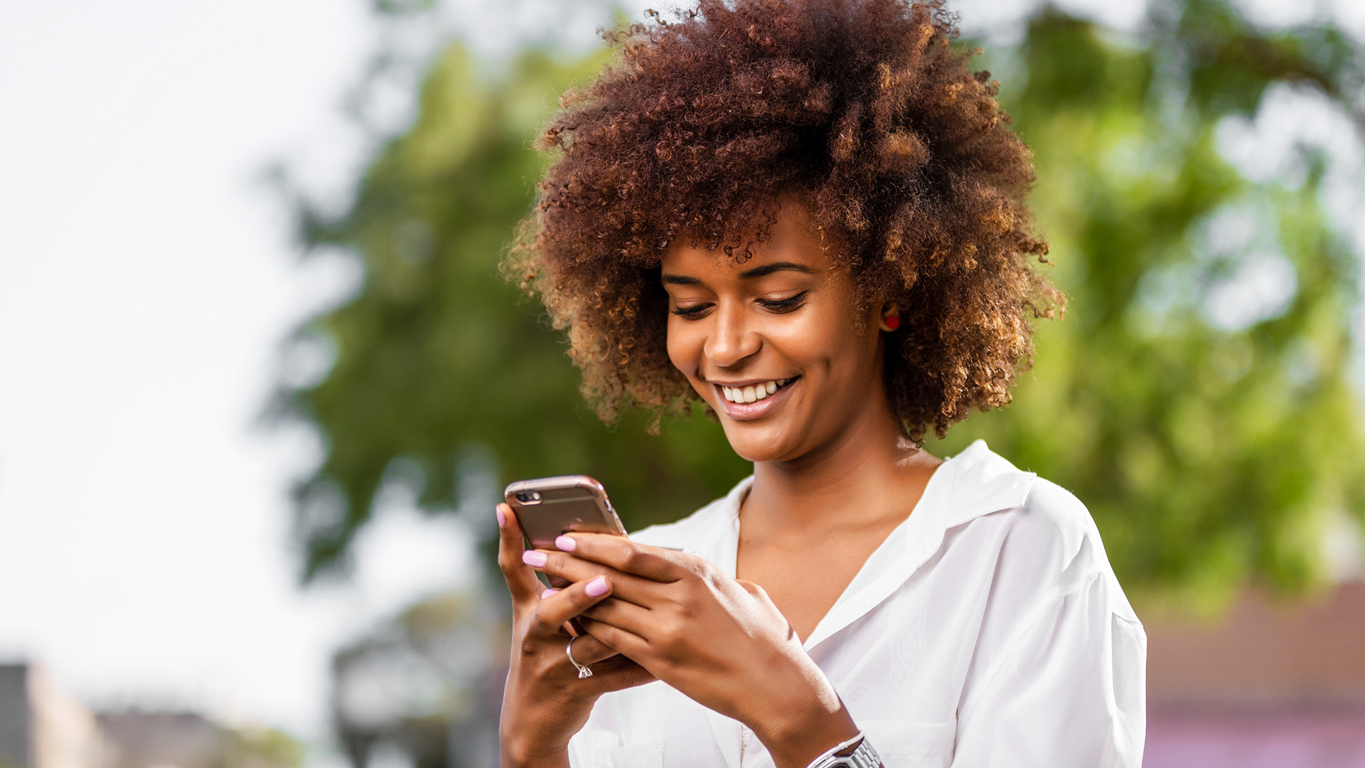Black woman wearing a white button down and her hair in an afro smiles while holding and looking at cellphone.