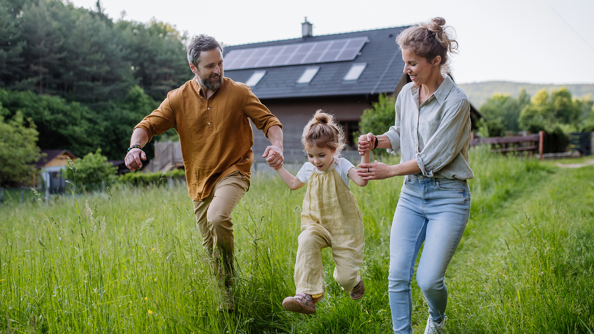 Two parents walk with their child in between them. They are smiling swinging the child by her arms. Behind them is a home with solar panels on the roof.