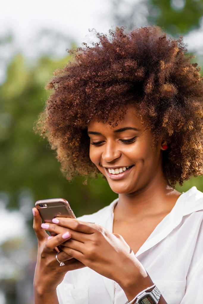 Black woman wearing a white button down and her hair in an afro smiles while holding and looking at cellphone.