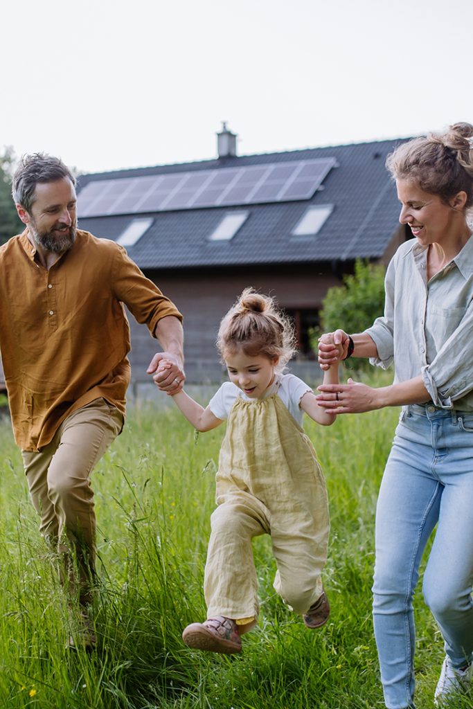 Two parents walk with their child in between them. They are smiling swinging the child by her arms. Behind them is a home with solar panels on the roof.