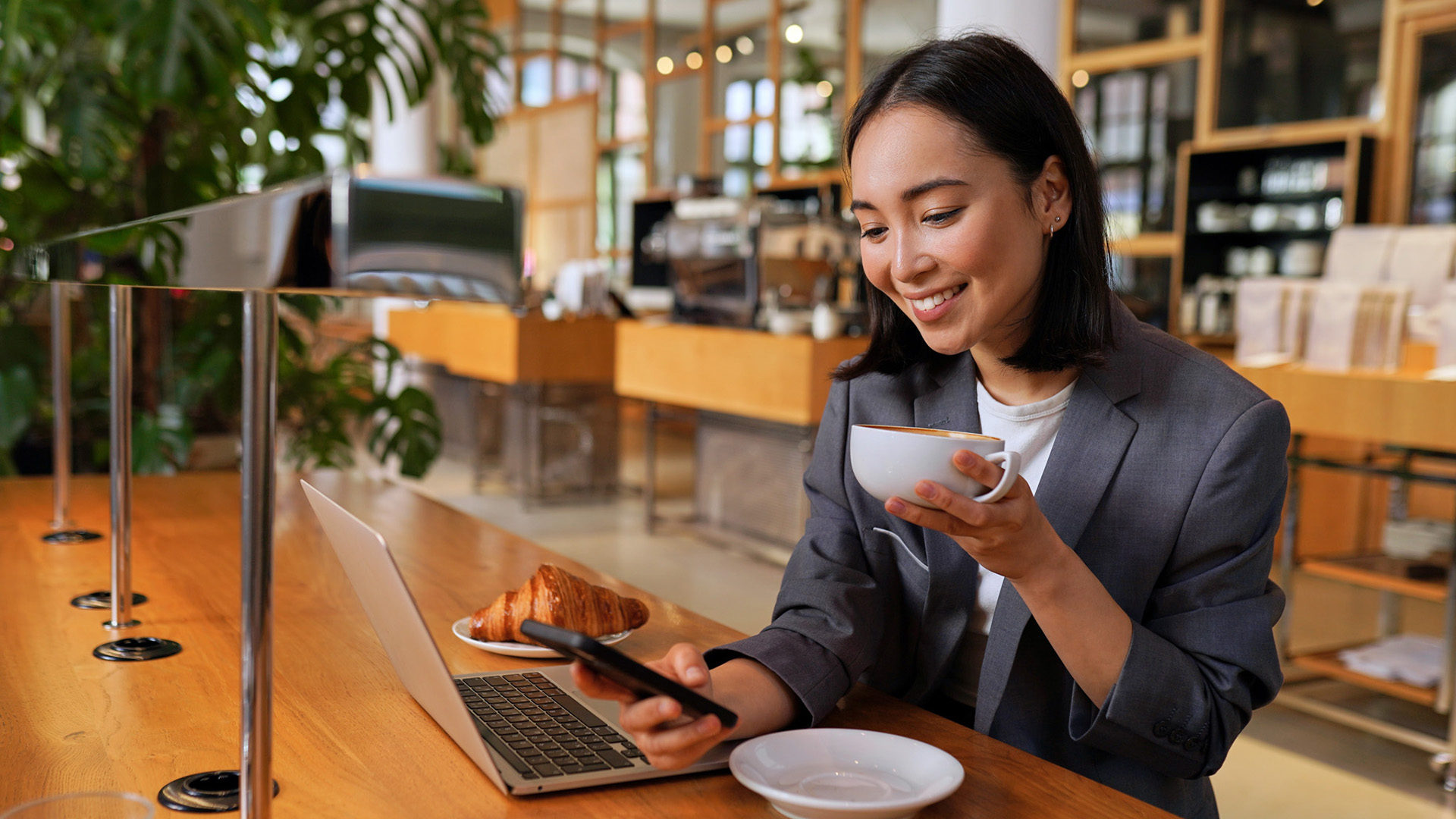 Young Asian business woman wearing suit drinking coffee using smartphone in cafe.