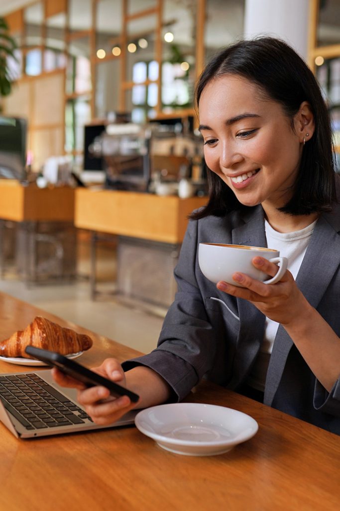 Young Asian business woman wearing suit drinking coffee using smartphone in cafe.