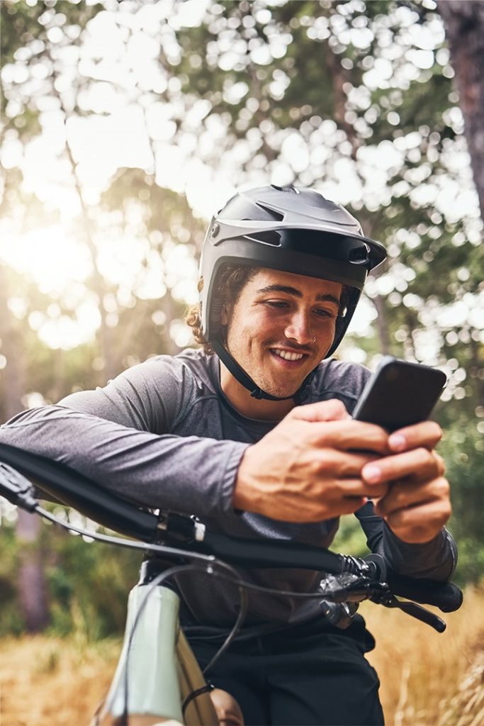 A man sitting on a bicycle wearing a helmet and smiling while he uses a smartphone.