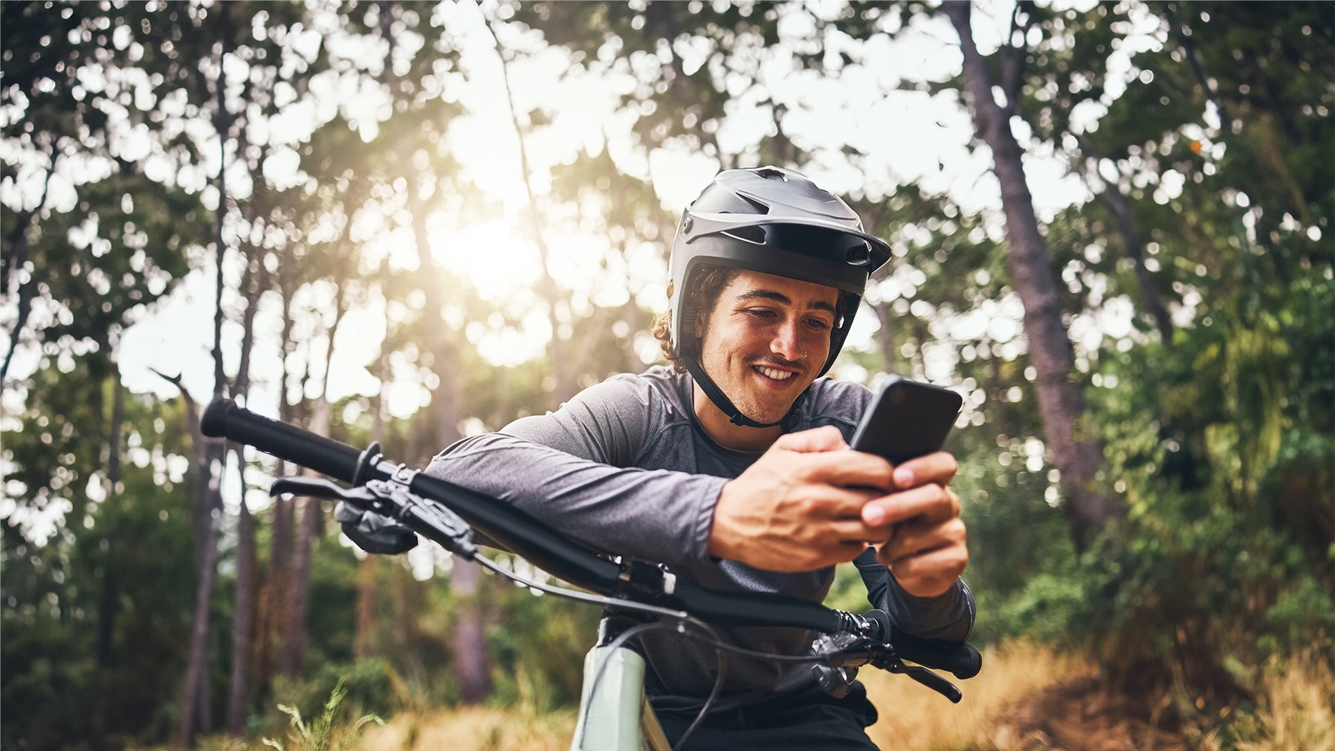 A man sitting on a bicycle wearing a helmet and smiling while he uses a smartphone.