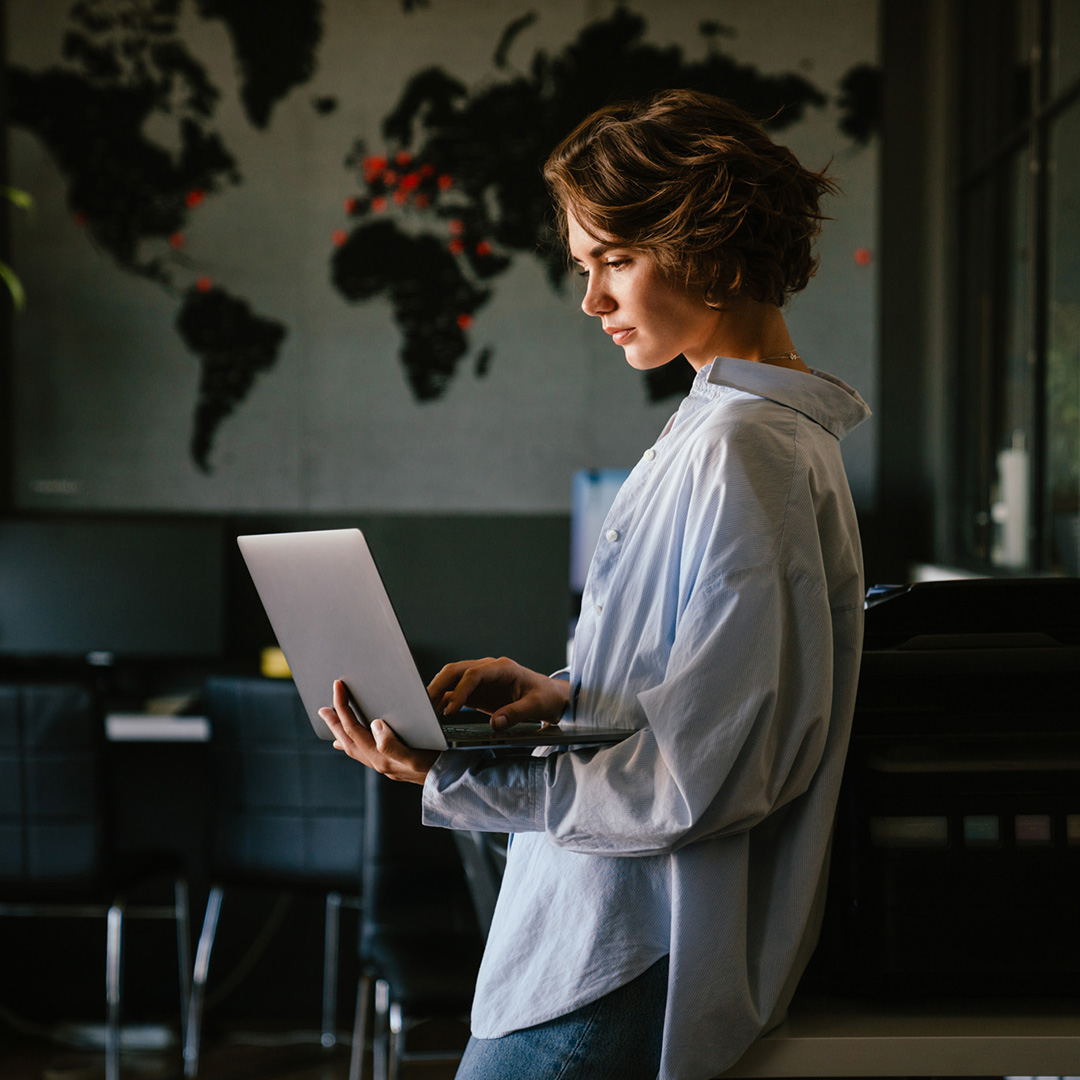 White woman with cropped hair in oversized button down shirt stands and holds a laptop computer while she works.