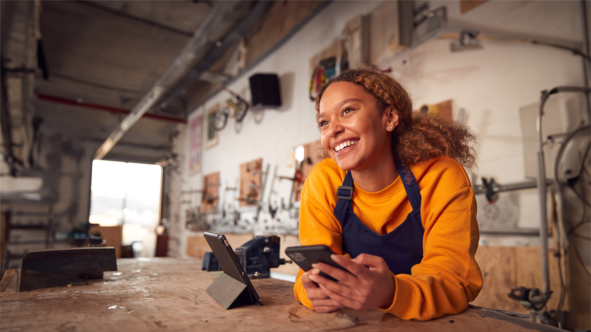 Young black woman with her natural hair in a ponytail leans on a counter with her cell phone in her hands. She is smiling and wearing an apron and orange shirt.