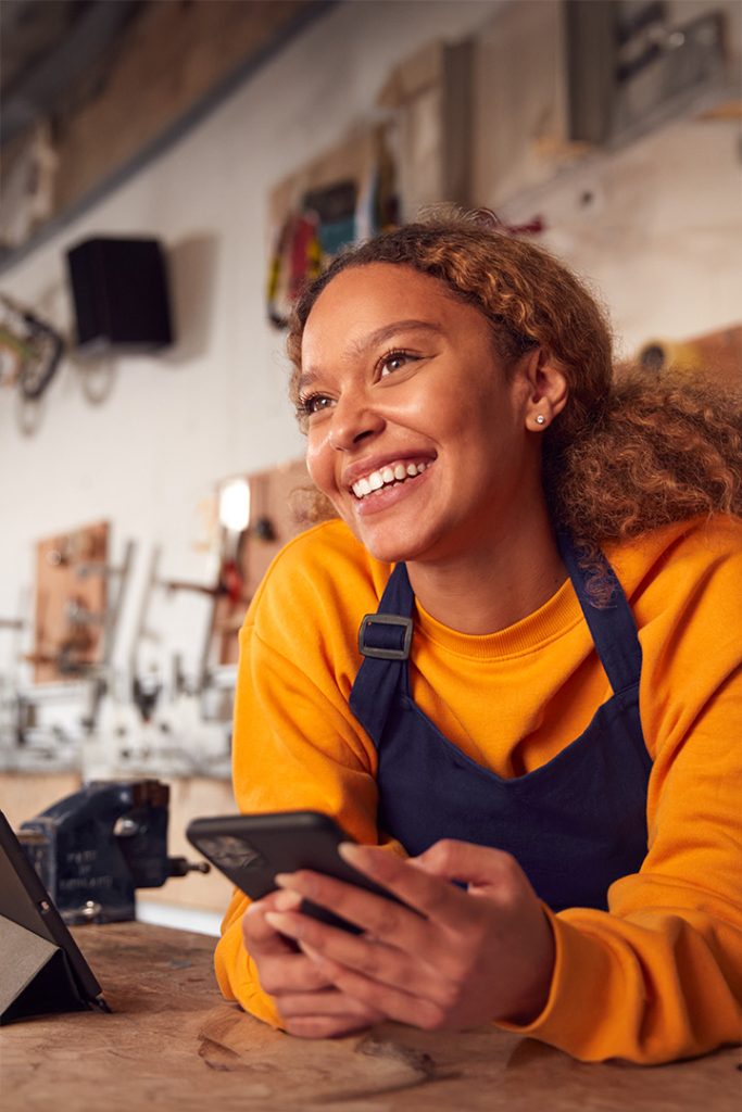 Young black woman with her natural hair in a ponytail leans on a counter with her cell phone in her hands. She is smiling and wearing an apron and orange shirt.