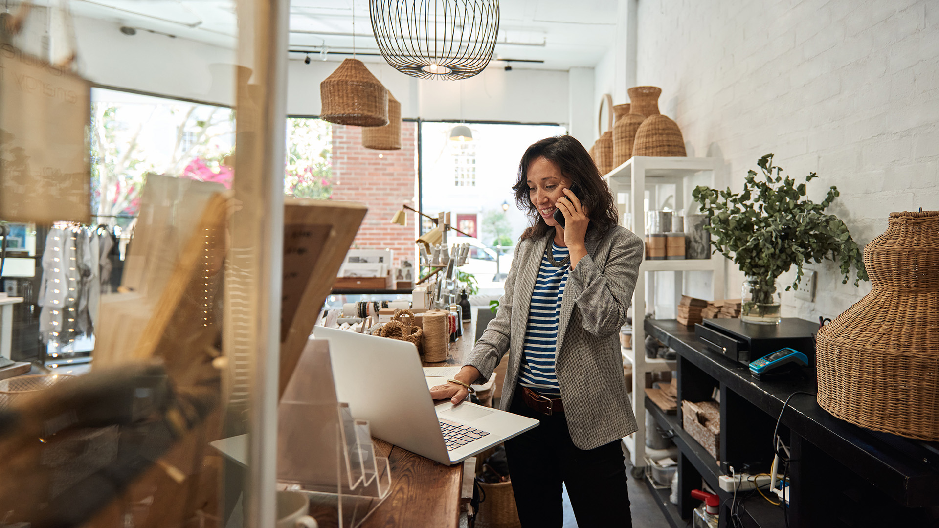 A woman standing running a small business. She has dark hair that she is wearing down around her shoulders and is wearing a striped t-shirt, blazer, and slacks. She is talking on the phone and using a laptop computer.