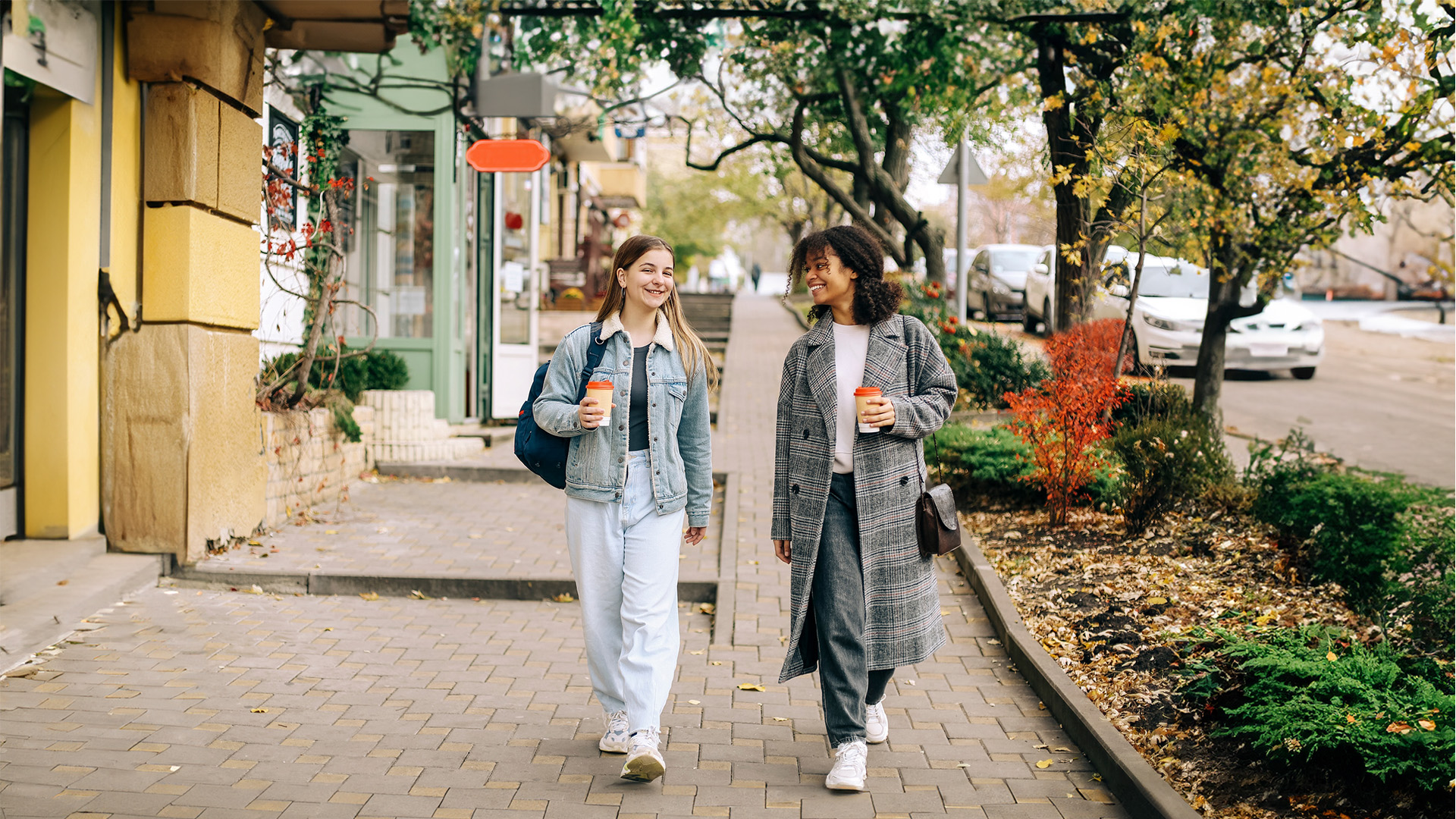 A young white woman and a young Black woman dressed for cool weather and walking down a street lined with retail shops. They are smiling and each holding a coffee.