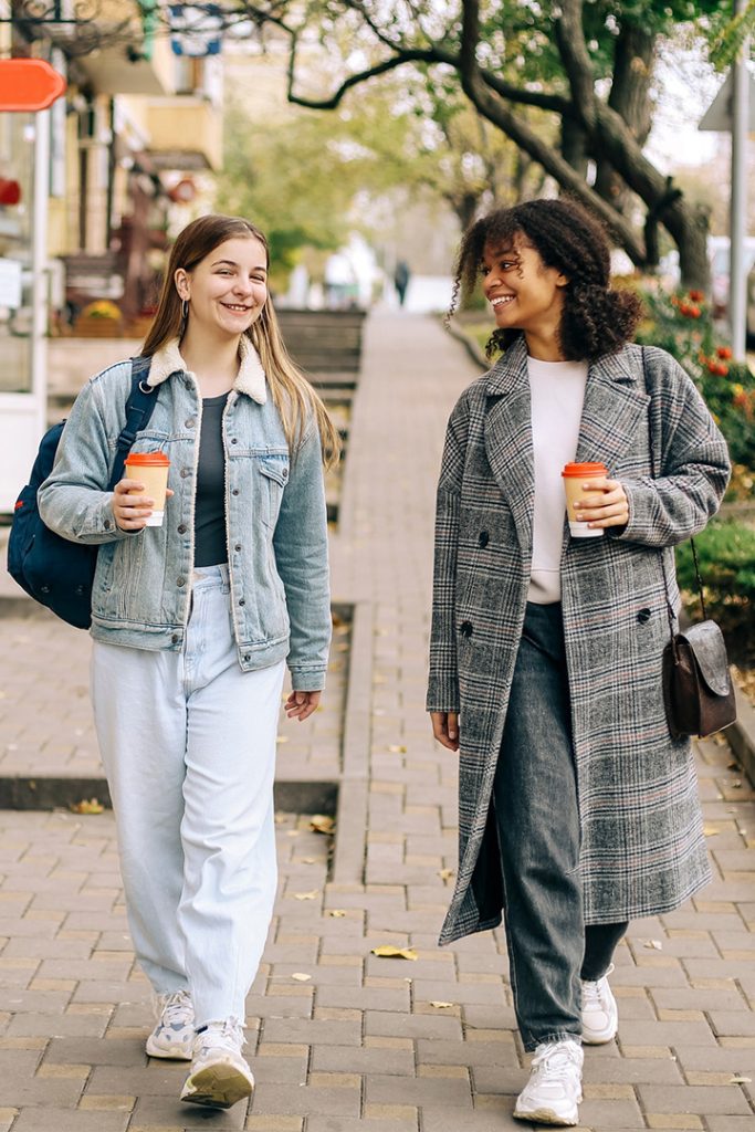 A young white woman and a young Black woman dressed for cool weather and walking down a street lined with retail shops. They are smiling and each holding a coffee.