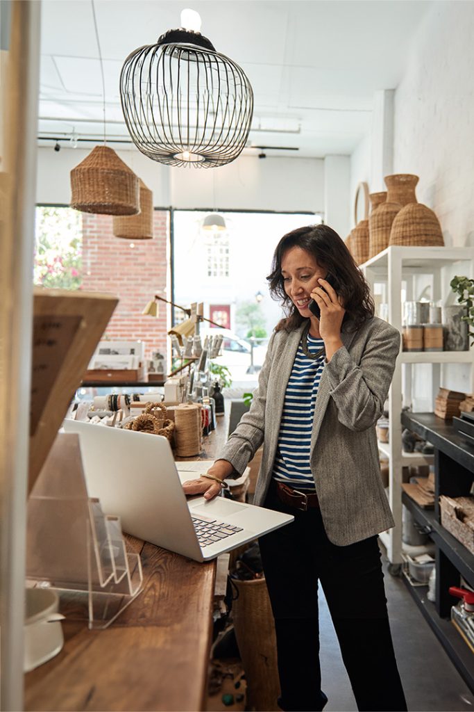A woman standing running a small business. She has dark hair that she is wearing down around her shoulders and is wearing a striped t-shirt, blazer, and slacks. She is talking on the phone and using a laptop computer.
