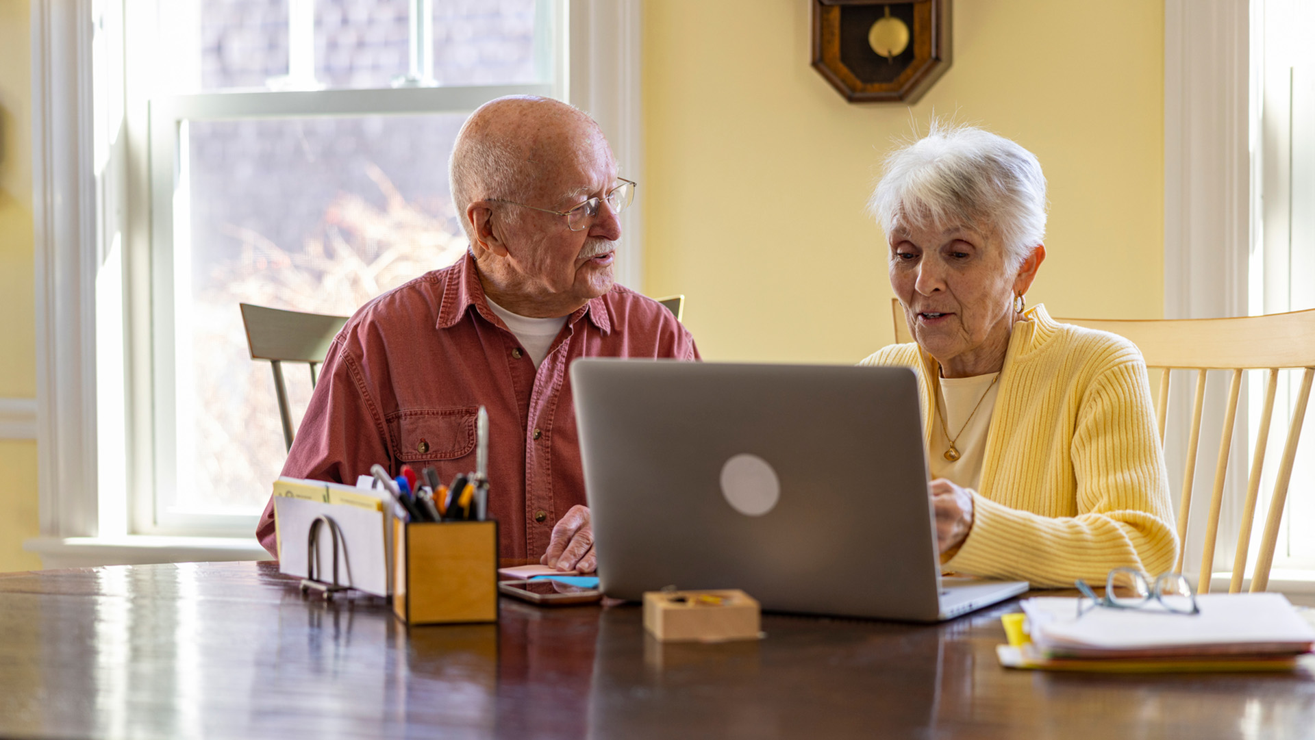 An elderly white couple sit at a dining table and use a laptop computer.