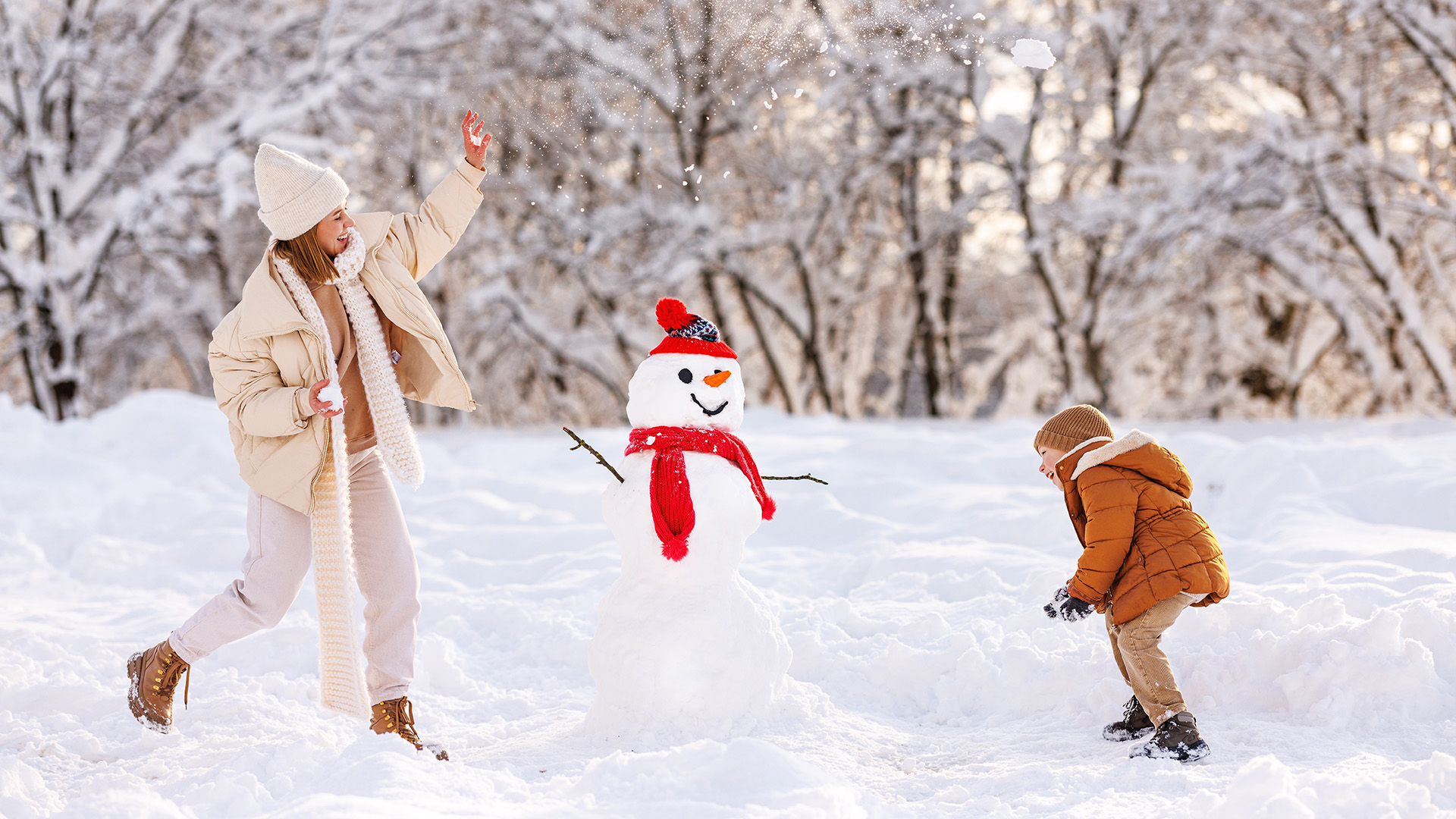 A young woman and child playing in the snow around a snowman.