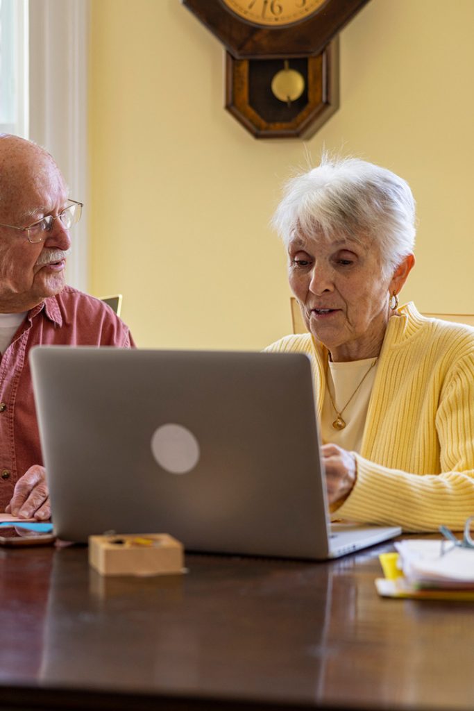 An elderly white couple sit at a dining table and use a laptop computer.