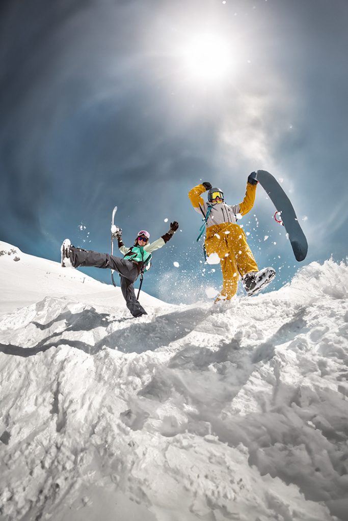 Two friends on a ski slope jumping in the snow. both are wearing heat to toe ski attire.