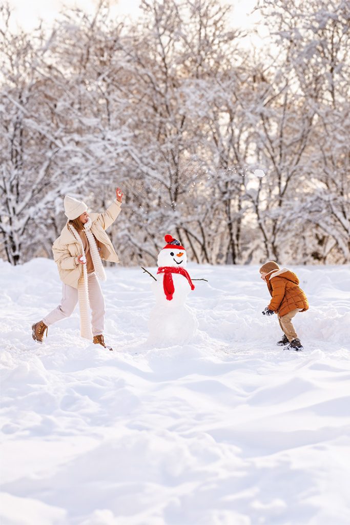 A young woman and child playing in the snow around a snowman.