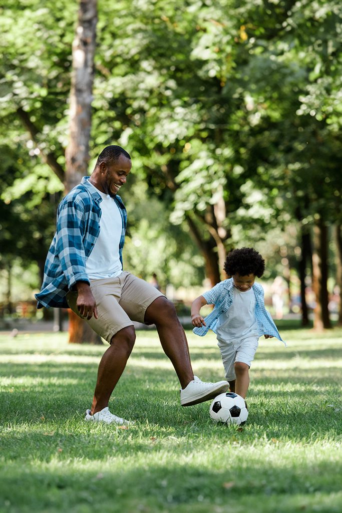 A Black man and his son play soccer in a grassy field.
