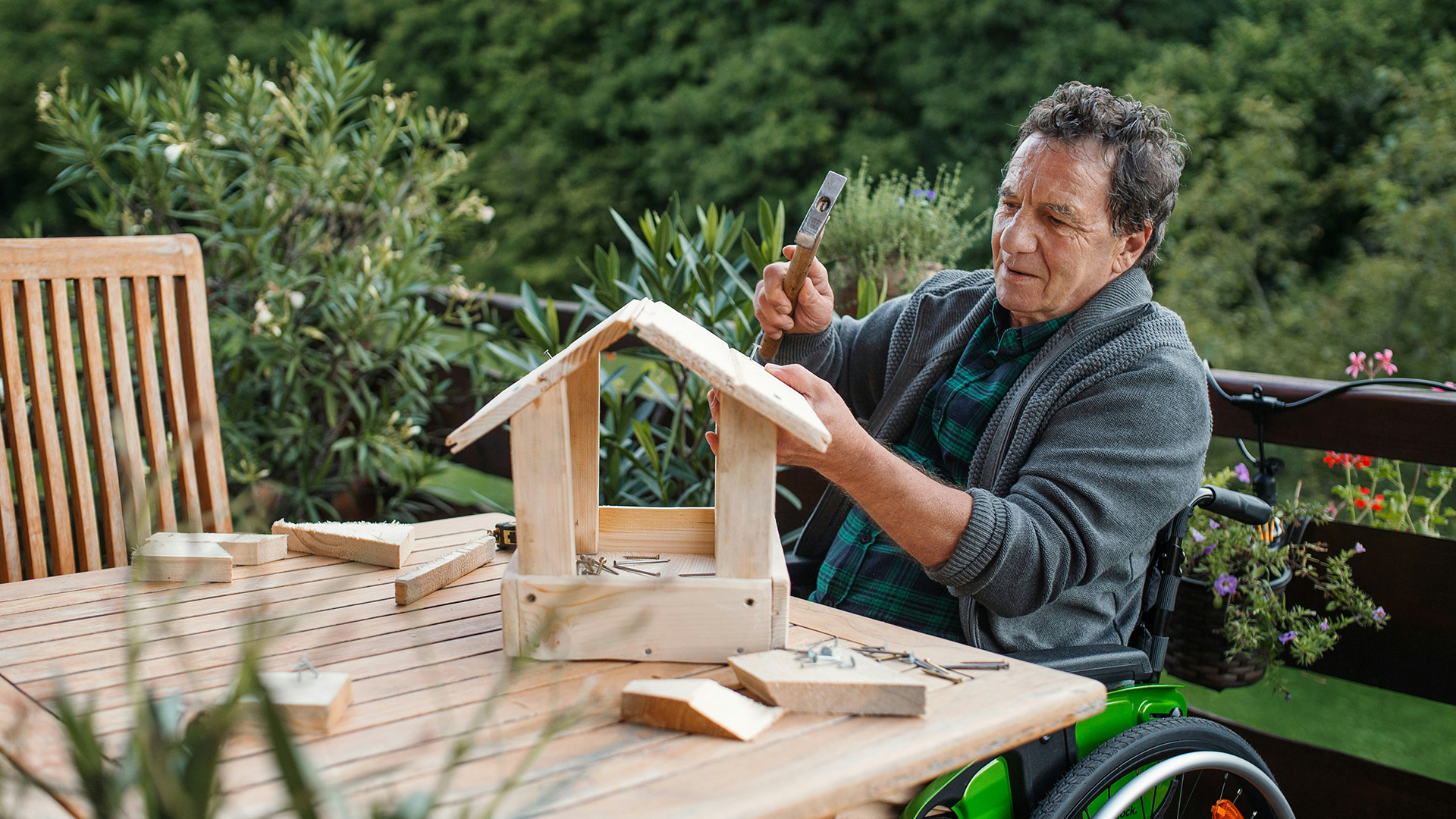 Older man in a wheelchair sitting at a table and building a bird feeder with a hammer.