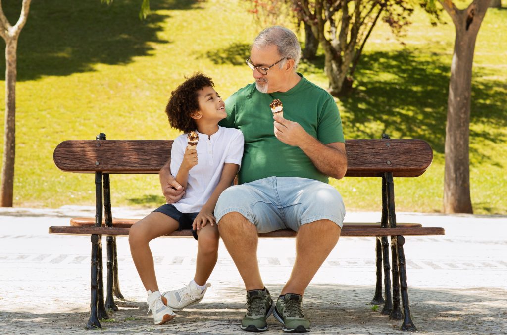 Older man eating ice cream in park on bench with younger child on nice, sunny day